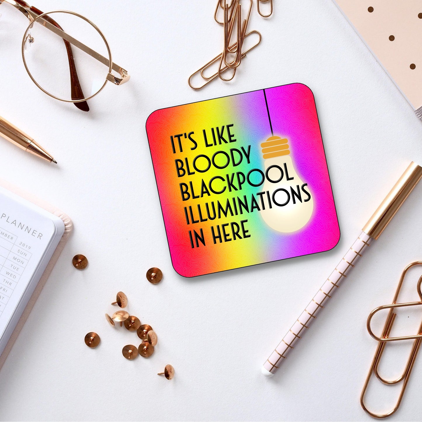 A colorful square coaster with rounded edges featuring text and a light bulb graphic. The background has a gradient of rainbow colors and the text reads 'IT'S LIKE BLOODY BLACKPOOL ILLUMINATIONS IN HERE'.