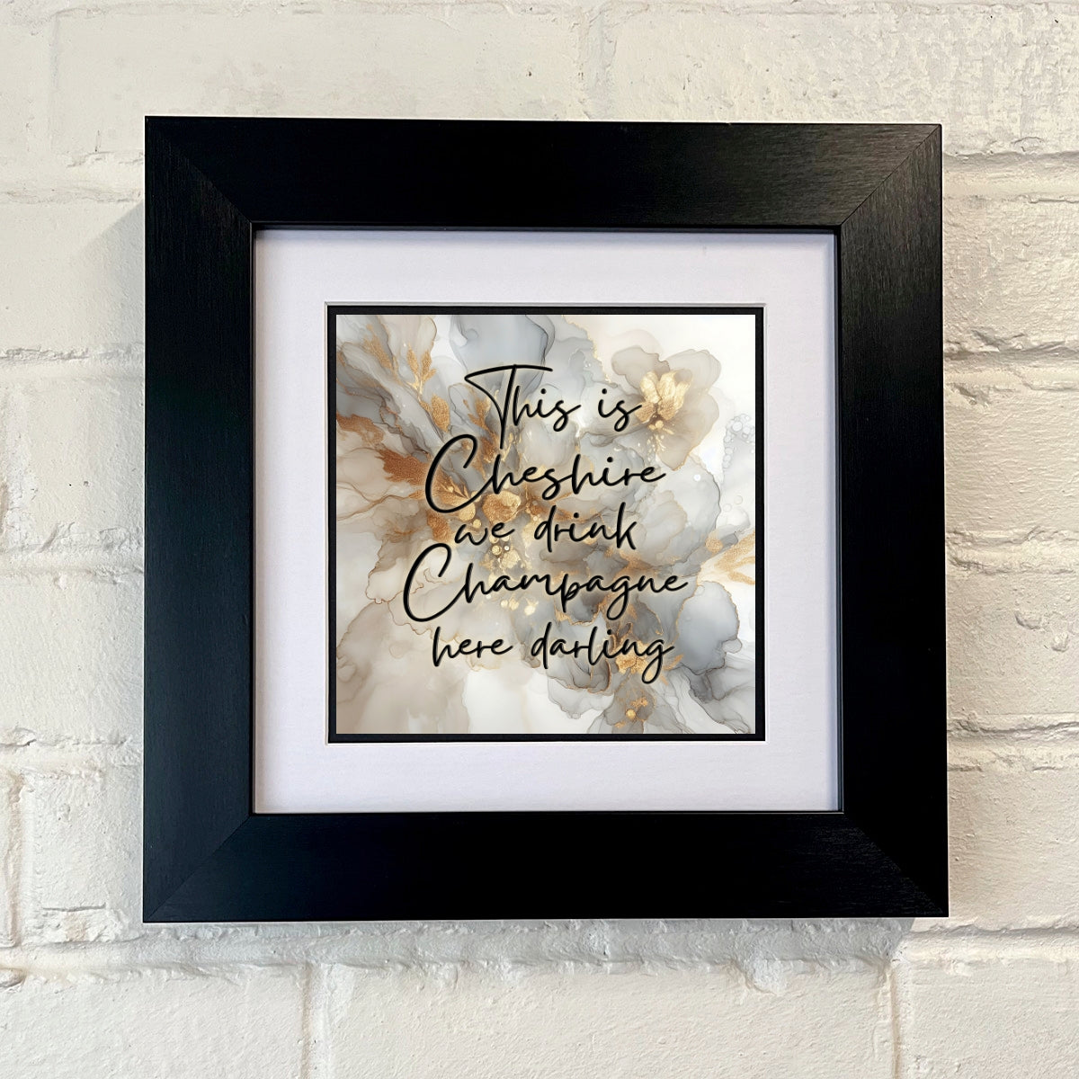 A framed print with the text 'This is Cheshire we drink Champagne here darling' displayed on a white wall.