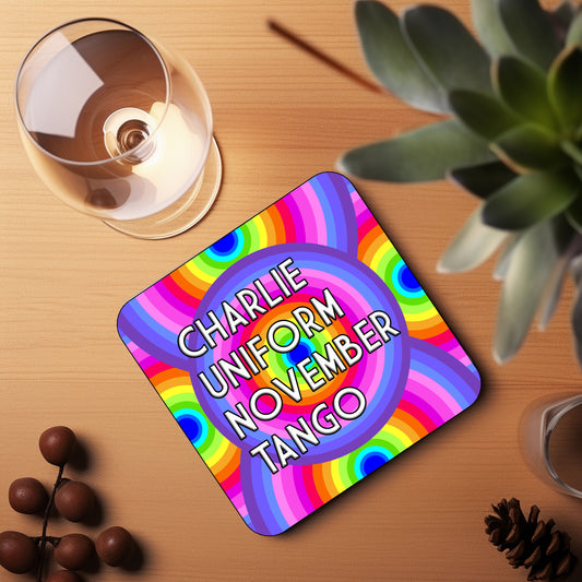 A colorful coaster with text 'CHARLIE UNIFORM NOVEMBER TANGO' displayed on a wooden surface with a glass of wine and a plant in the background.