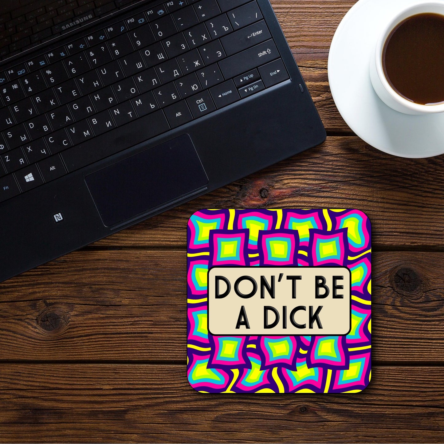 Colorful coaster with 'DON'T BE A DICK' text on a wooden desk with a laptop and coffee.