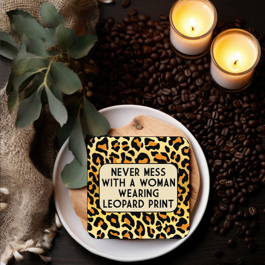 A square coaster with leopard print design and the text 'NEVER MESS WITH A WOMAN WEARING LEOPARD PRINT' displayed on a plate with coffee beans and candles in the background.
