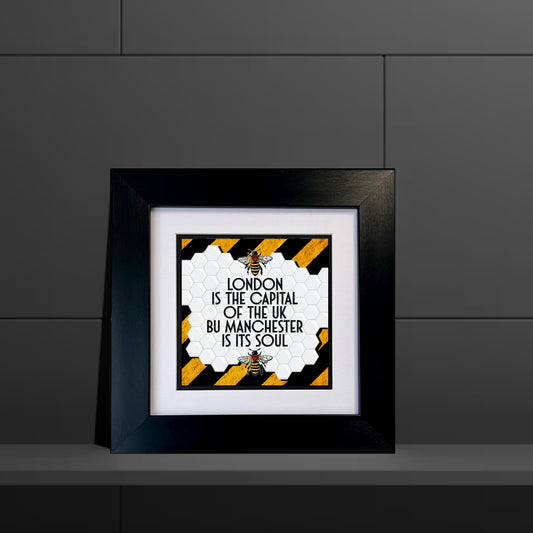 A black square frame with a print that states 'LONDON IS THE CAPITAL OF THE UK BUT MANCHESTER IS ITS SOUL' with a Manchester bee logo and yellow and black stripes.