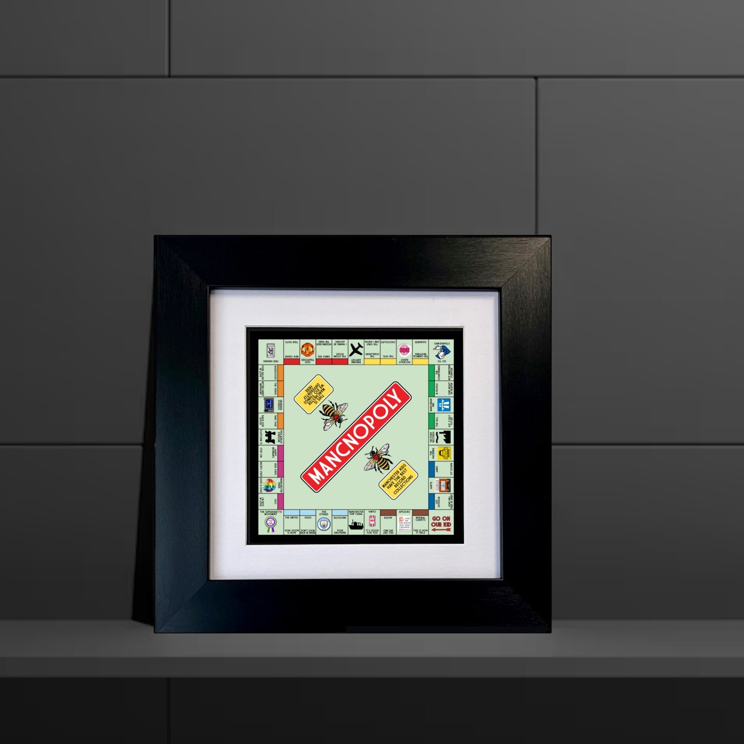 A framed print of a Manchester-themed Monopoly board game, with a chunky black frame, displayed against a white wall.