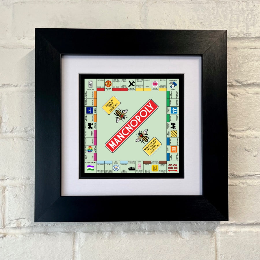 A framed print of a Manchester-themed Monopoly board game, with a chunky black frame, displayed against a white wall. SU MACE GIFTS