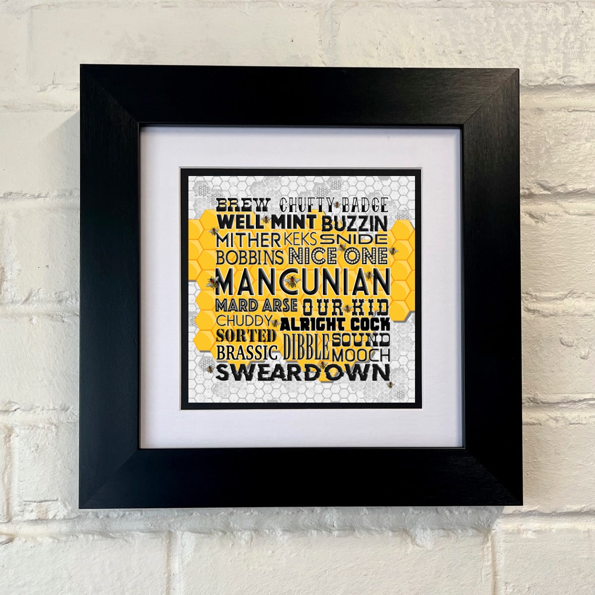 A framed print with black and yellow text, containing Manchester slang, displayed against a white brick wall.
