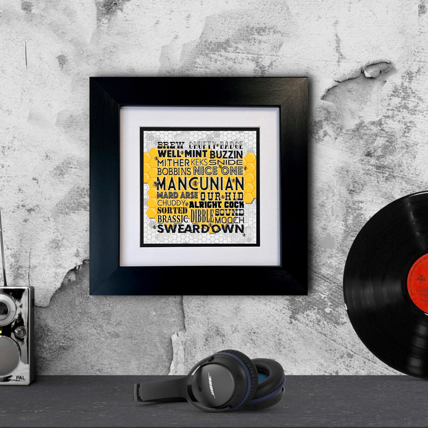 A framed print with black and yellow text, containing Manchester slang, displayed against a white brick wall.