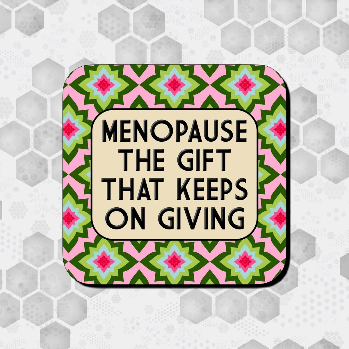 Coaster with a pink background featuring a repeating geometric floral pattern in green, red, and blue, overlaid with a rounded beige box containing bold black text that reads ‘Menopause – the gift that keeps on giving