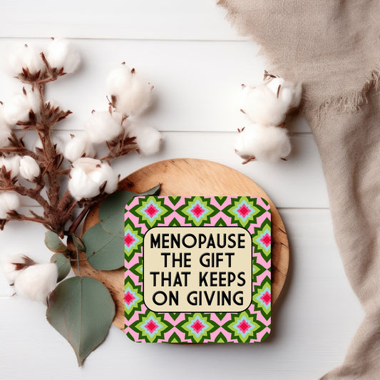 Coaster with a pink background featuring a repeating geometric floral pattern in green, red, and blue, overlaid with a rounded beige box containing bold black text that reads ‘Menopause – the gift that keeps on giving