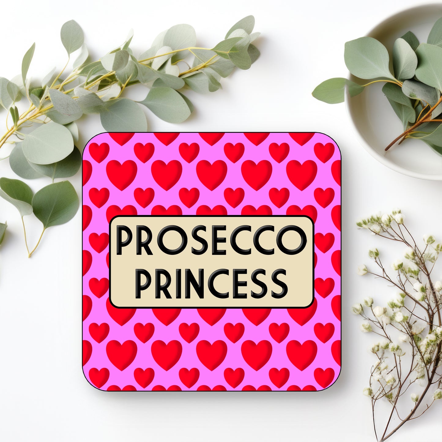 Coaster with 'Prosecco Princess' text on a pink background with red hearts, surrounded by greenery.