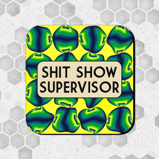 Coaster with a bold yellow background covered in abstract green and blue geometric shapes, featuring a rounded beige box in the center with bold black text reading ‘Shit Show Supervisor.