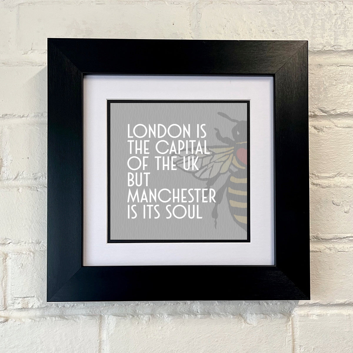 A black square frame with a print that states 'LONDON IS THE CAPITAL OF THE UK BUT MANCHESTER IS ITS SOUL' with a Manchester bee logo and GREY BACKGROUND
