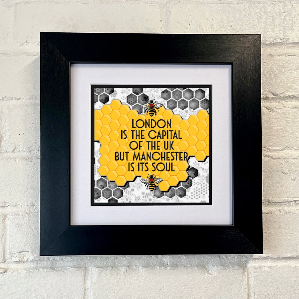 A black square frame with a print that states 'LONDON IS THE CAPITAL OF THE UK BUT MANCHESTER IS ITS SOUL' with a Manchester bee logo and yellow and GREY HONEYCOMB PATTERN