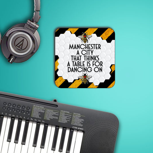 Coaster design with a white honeycomb background and black-and-yellow hazard stripes, featuring the Manchester worker bee above and below bold text that reads ‘Manchester, a city that thinks a table is for dancing on