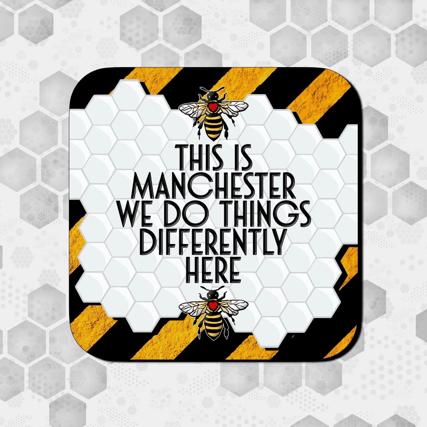 Coaster featuring a white honeycomb pattern with bold text reading ‘This is Manchester, we do things differently here,’ framed by two Manchester worker bees and black-and-yellow hazard stripes in the background