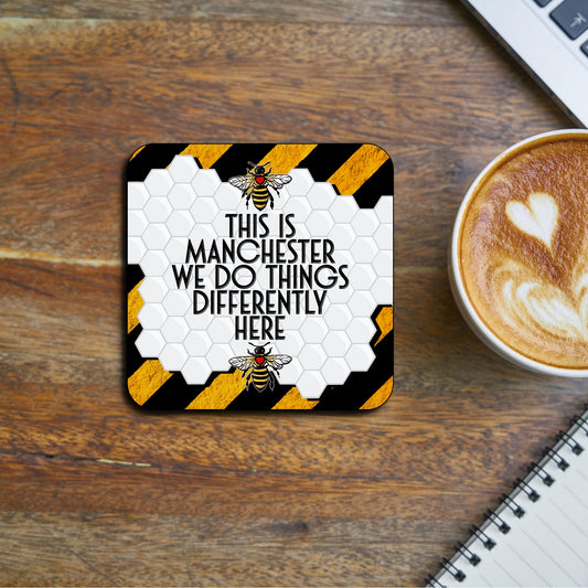 Coaster featuring a white honeycomb pattern with bold text reading ‘This is Manchester, we do things differently here,’ framed by two Manchester worker bees and black-and-yellow hazard stripes in the background