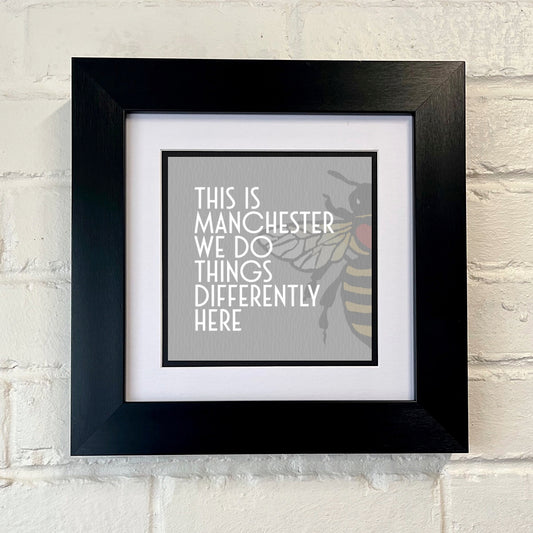 A framed print with a black border containing a graphic that says 'THIS IS MANCHESTER WE DO THINGS DIFFERENTLY HERE' with a Manchester bee and a GREY patterned background.