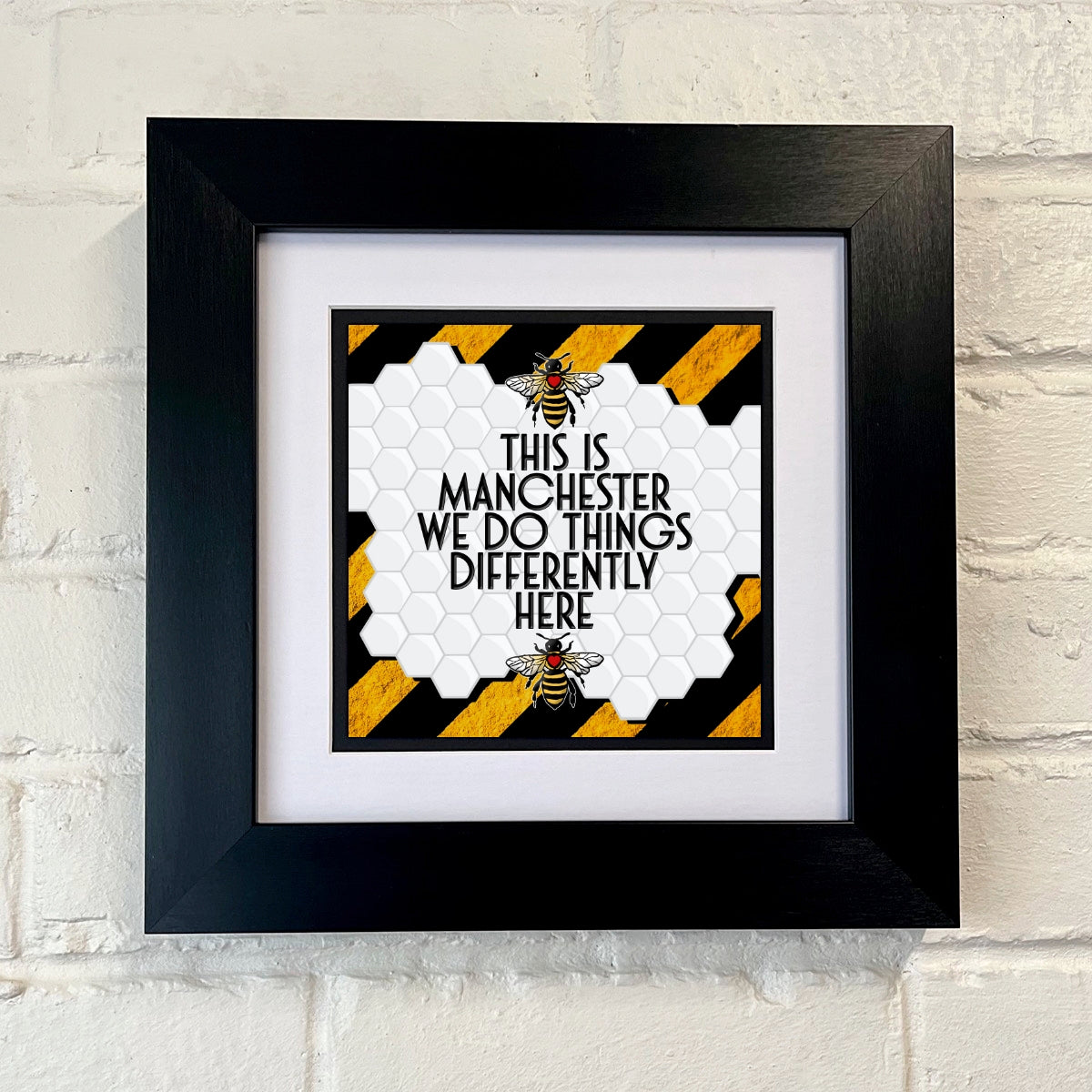 A framed print with a black border containing a graphic that says 'THIS IS MANCHESTER WE DO THINGS DIFFERENTLY HERE' with a Manchester bee and a STRIPE patterned background.