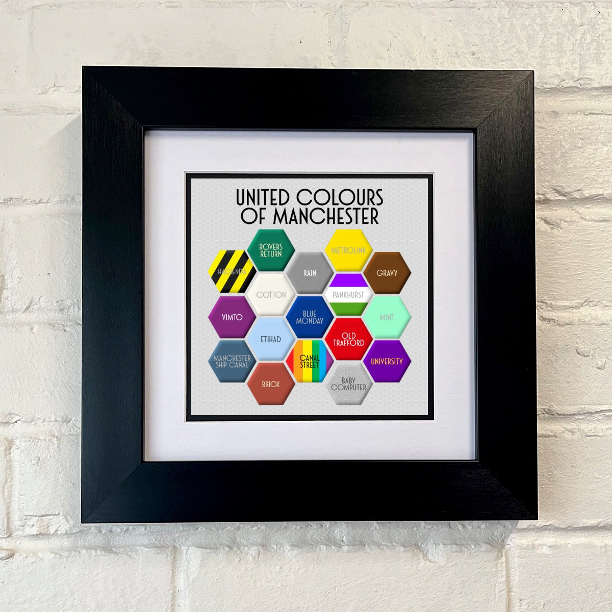 A colourful FRAMED print titled 'UNITED COLOURS OF MANCHESTER', featuring a honeycomb pattern with different colours and text elements representing various aspects of Manchester.SU MACE GIFTS