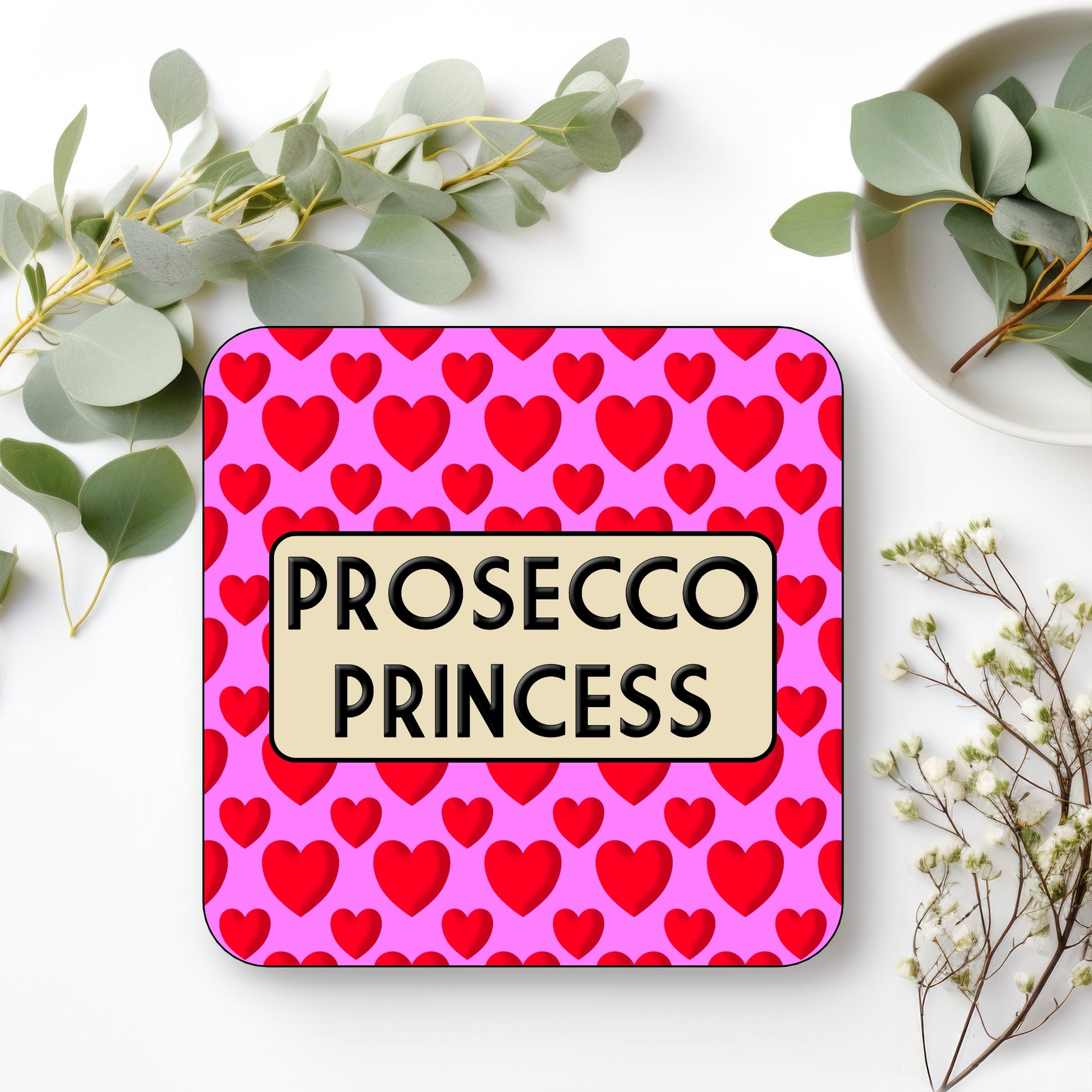 Coaster with 'Prosecco Princess' text on a pink background with red hearts, surrounded by greenery.