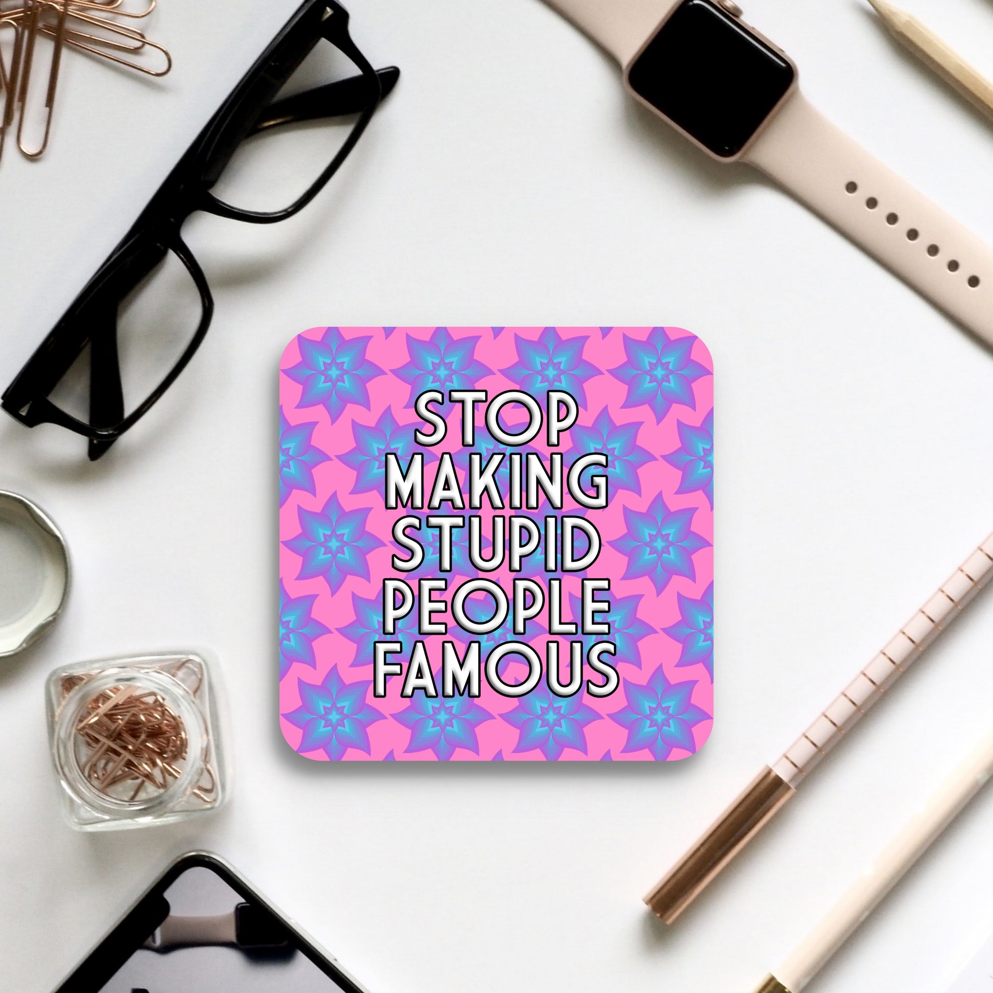 Coaster with text 'Stop making stupid people famous' on a pink and blue patterned background, surrounded by office supplies.