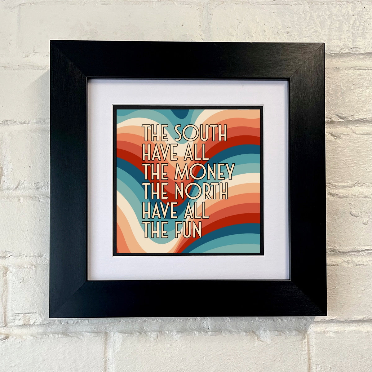 A framed print with the text 'THE SOUTH HAVE ALL THE MONEY THE NORTH HAVE ALL THE FUN' on a colorful background, displayed against a white brick wall.